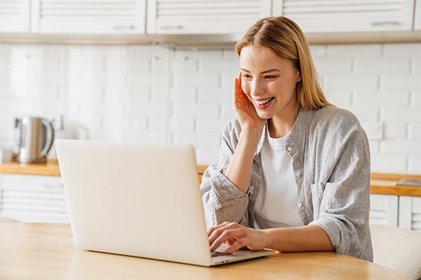 Joyful-blonde-girl-smiling-and-using-laptop-while-sitting-at-home-kitchen